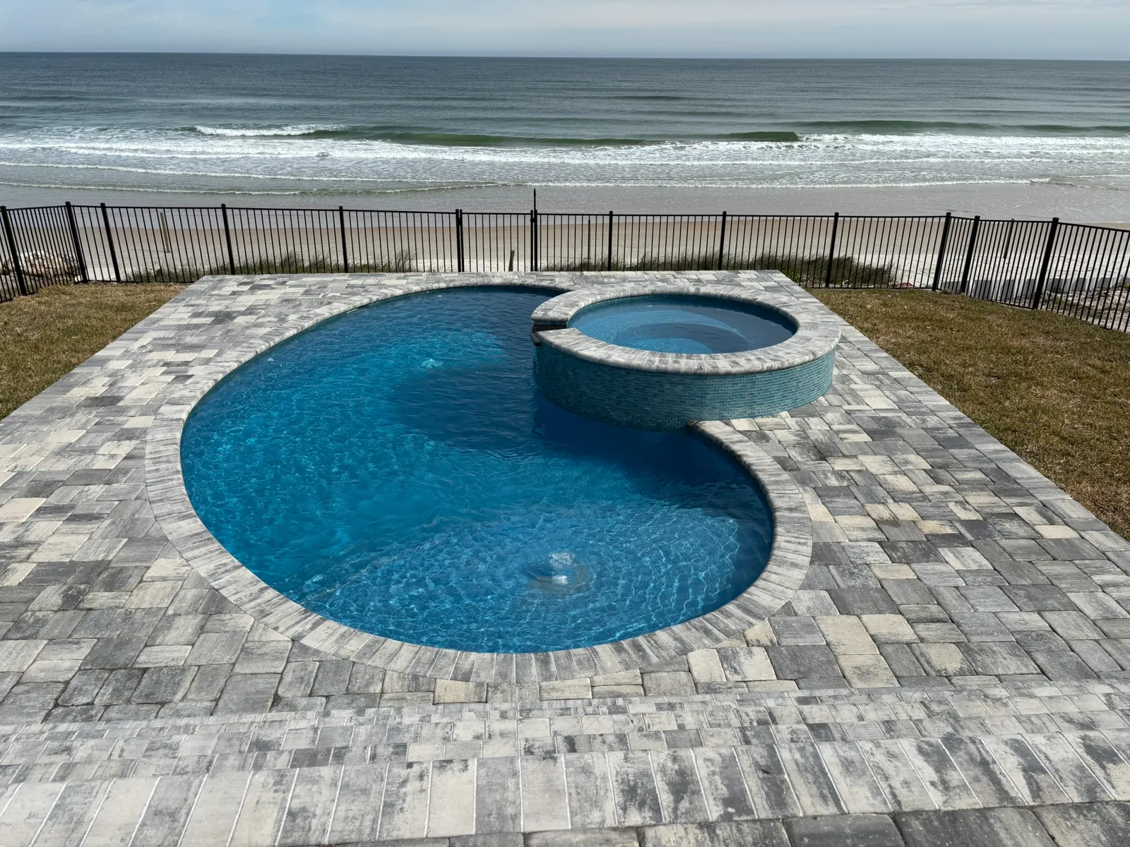 Oceanfront freeform pool with round raised spa and gray paver deck overlooking the beach