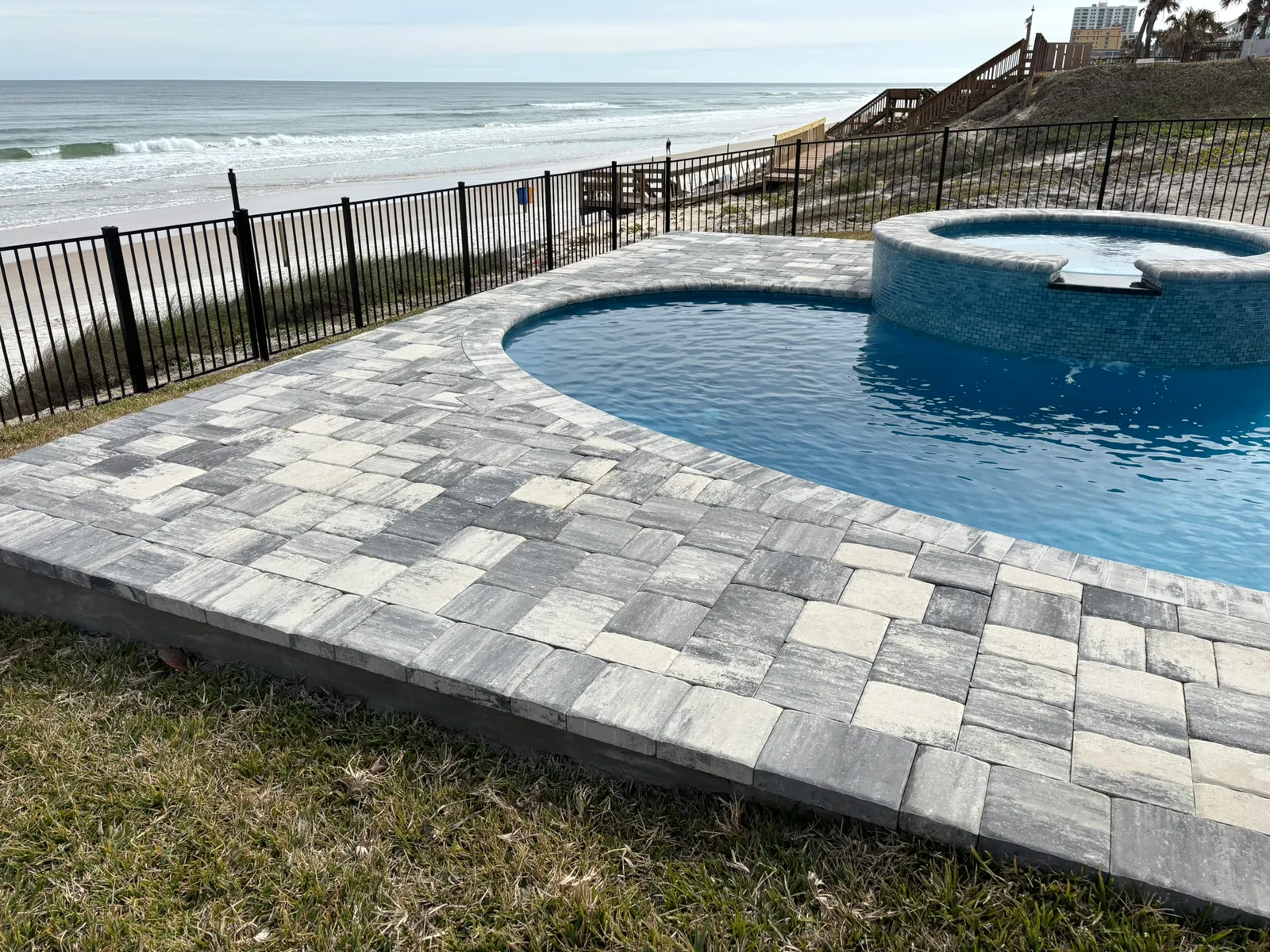 Freeform pool and spa with a wide gray paver deck beside the dunes and beach